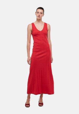 Femme debout portant une robe rouge sans manches, longue jusqu'au sol, avec un décolleté en V et des talons rouges ouverts sur un fond clair uni.