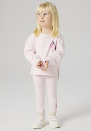 Niña rubia con gafas que lleva una sudadera color rosa pálido con fresas y leggings, con zapatillas blancas, de pie frente a un fondo liso.