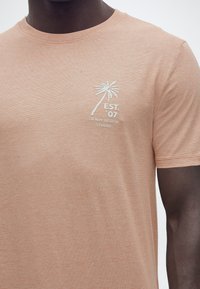 Ανοιχτό καφέ μπλουζάκι με λευκό σχέδιο φοίνικα και κείμενο «EST. '07 DENIM DESIGN STUDIO» στο αριστερό στήθος, φορεμένο από άτομο με σκουρόχρωμο δέρμα.