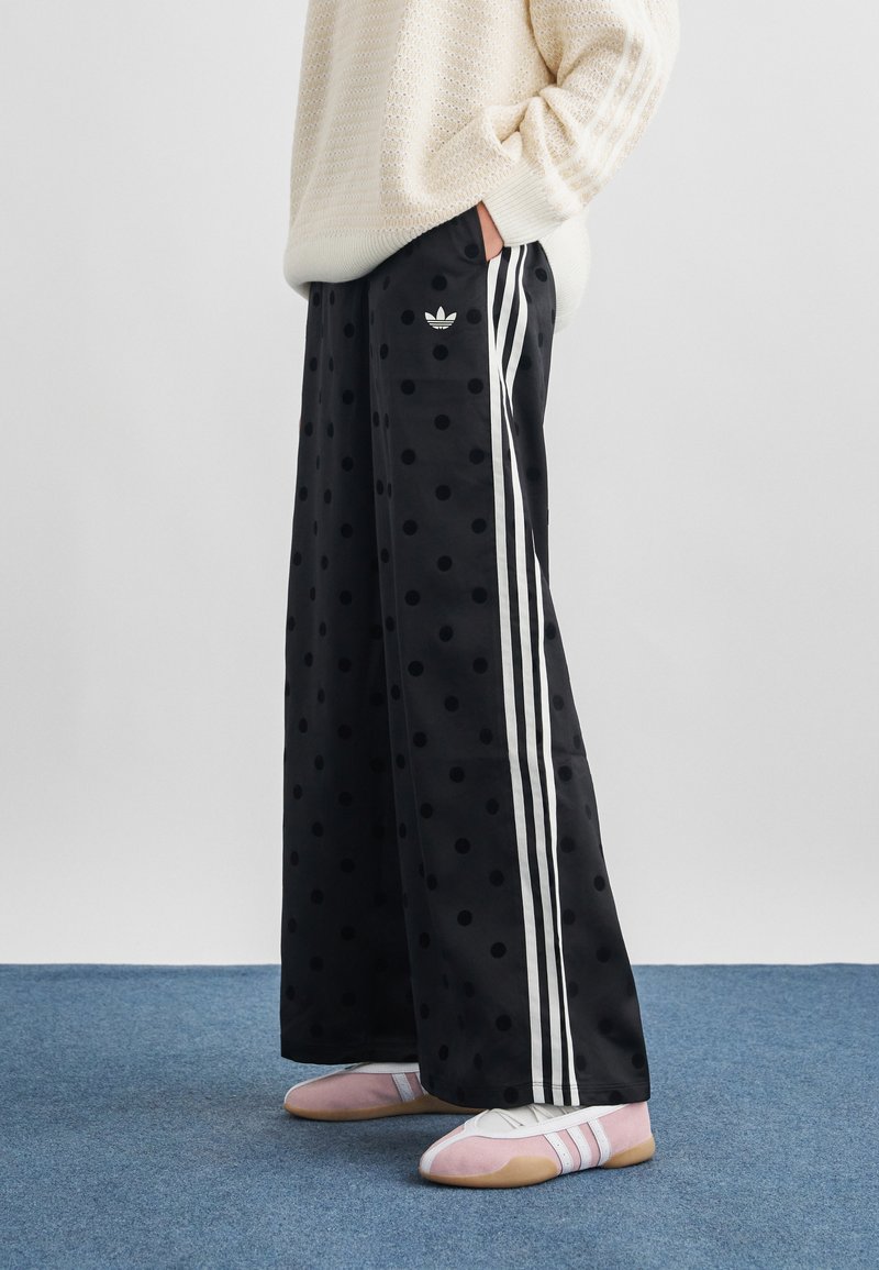 Άτομο που φοράει μαύρο παντελόνι με φαρδιά πόδια, μεγάλα πουά και λευκές ρίγες, ένα κρεμ πλεκτό πουλόβερ και ροζ και λευκά αθλητικά παπούτσια.