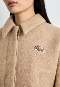 Manteau en laine beige avec un col à boutons, une poche unique et un logo de crocodile brodé en marron et rouge sur la zone du côté gauche de la poitrine.