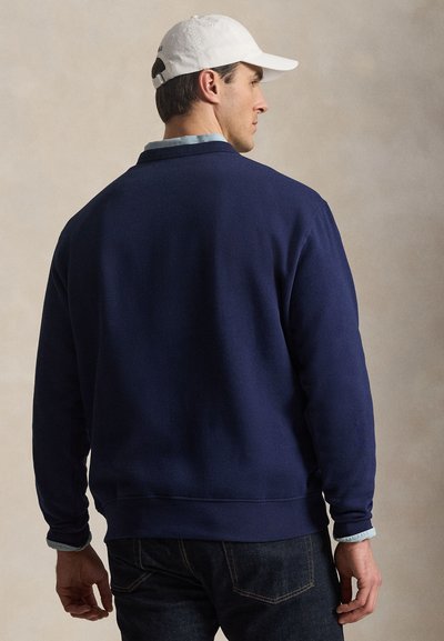 Άνδρας που φοράει μπλε ναυτικό πουλόβερ πάνω από ανοιχτό μπλε πουκάμισο με γιακά, σκούρο τζιν και λευκό καπέλο μπέιζμπολ, γυρισμένος με την πλάτη σε απλό φόντο.