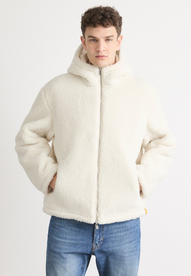 Bijela flis jakna sa zatvaračem i kapuljačom, s mekim materijalom i elastičnim manžetama. Uparena s plavim trapericama.