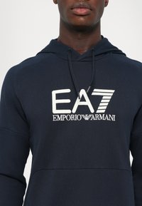 La felpa blu navy presenta un grande logo bianco EA7 Emporio Armani, cappuccio con cordino e un tessuto morbido e testurizzato. Design semplice con tasca a marsupio.