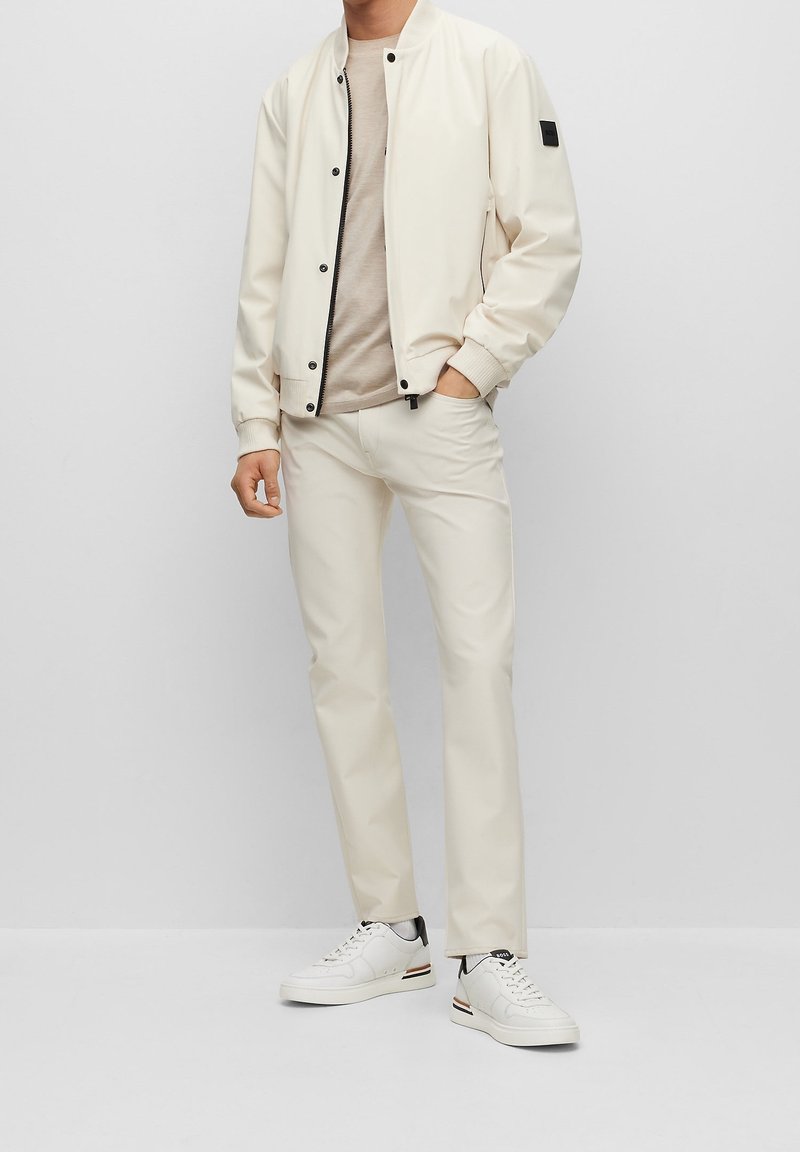 Vit lättvikts bomberjacka över en beige t-shirt, parat med krämfärgade slim-fit byxor och vita sneakers med svarta och gyllene detaljer.