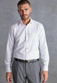 Weißes Hemd mit schmaler Passform, knöpfbarer Vorderseite und klassischem Kragen, kombiniert mit grauen strukturierten Hosen und einem schwarzen Gürtel.
