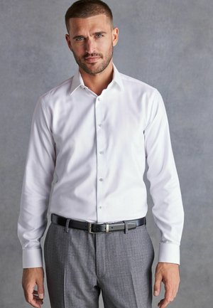 Weißes Hemd mit schmaler Passform, knöpfbarer Vorderseite und klassischem Kragen, kombiniert mit grauen strukturierten Hosen und einem schwarzen Gürtel.