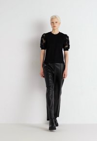 Svart topp med blondepuff ermer, kombinert med svarte skinnbukser. Glatt tekstur og tettsittende design. Modellen står mot en nøytral bakgrunn.