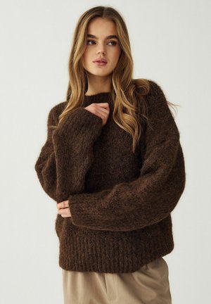 Jeune femme aux longs cheveux ondulés portant un pull en laine marron foncé à texture duveteuse et un pantalon beige, regardant sur le côté devant un fond uni.