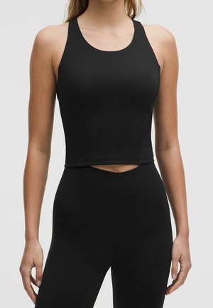 Woman wearing a fitted black sleeveless crop top and high-waisted black leggings, standing against a plain light background.