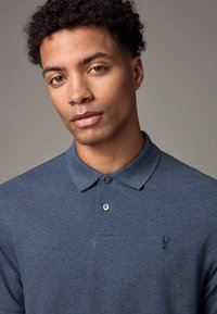 Camisa polo de tela azul oscuro. Presenta un cuello con solapa, una tapeta de dos botones y un pequeño logo bordado en el lado izquierdo del pecho.