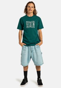 Camiseta de algodón verde con diseño gráfico blanco, combinada con pantalones cortos de mezclilla azul claro. El modelo lleva zapatillas negras y medias negras hasta la rodilla.