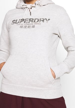 Lichtgrijze hoodie met de tekst "SUPERDRY Tokyo London Paris" en Japanse tekens, voorzak en trekkoordkap, gedragen door een persoon met handen in de zakken.