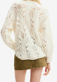 Sweter w kolorze kremowym, wykonany na drutach z skomplikowanym wzorem warkoczy i otwartym splotem, w połączeniu z zieloną spódnicą. Faktura wydaje się lekka i przewiewna.