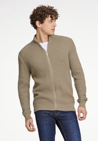 Suéter de punto con cremallera en marrón claro con un patrón de diamantes texturizado y puños acanalados; combinado con jeans azul oscuro.