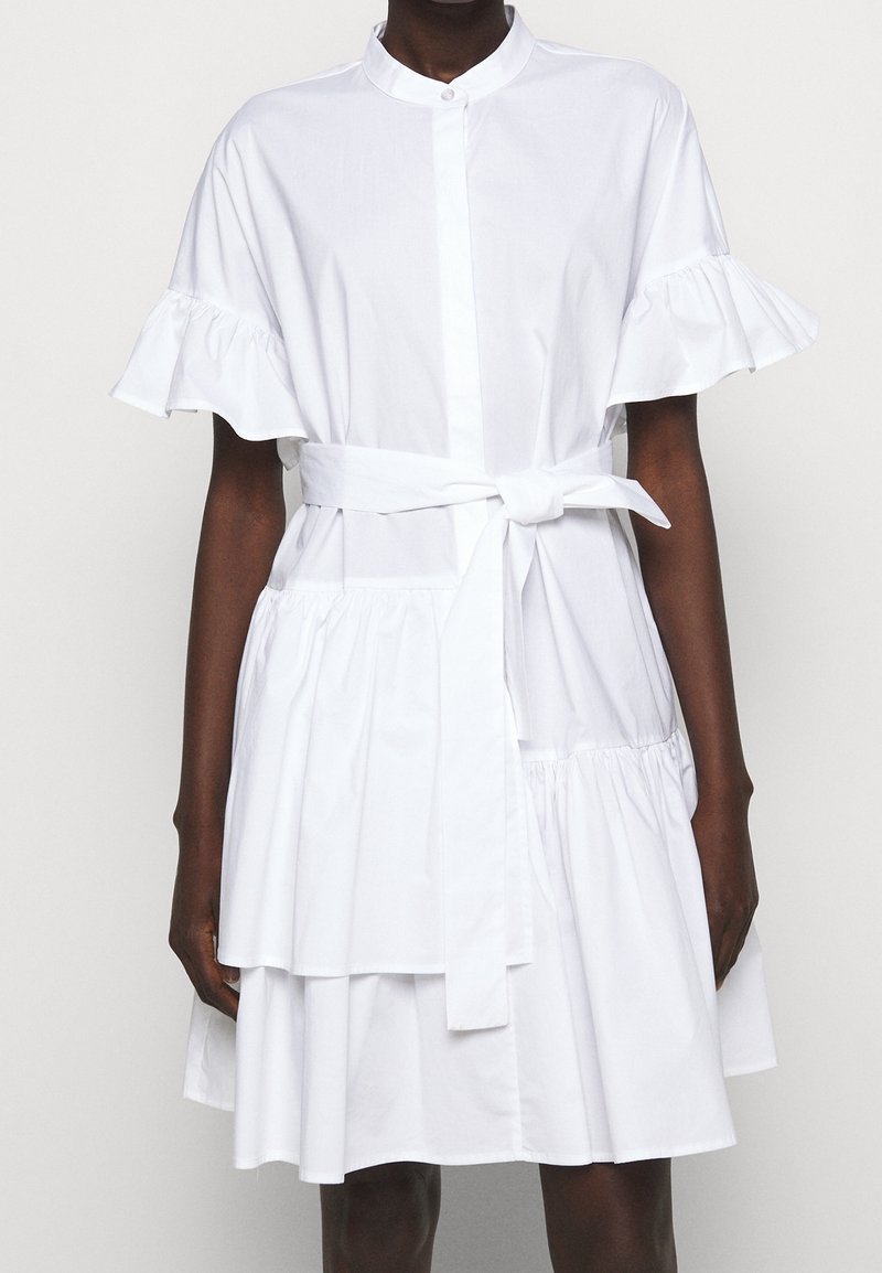 Robe blanche à étages avec manches courtes volantées, col montant, ouverture boutonnée sur le devant et ceinture nouée à la taille.