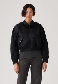 Γυναίκα με κοντά σκούρα μαλλιά που φοράει μαύρο μπουφάν bomber πάνω από σκούρο τοπ και σκούρο τζιν, στέκεται μπροστά σε απλό ανοιχτό φόντο.