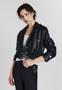Schwarze, kurz geschnittene Lederjacke mit silbernen Druckknöpfen, Reißverschluss-Taschen und elastischem Bund. Über einem weißen Oberteil getragen.