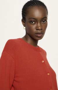 Frau mit geflochtenen Haaren, die kleine goldene Creolen und eine rote, geknöpfte Strickjacke trägt, die nach vorne blickt und einen neutralen Gesichtsausdruck hat.
