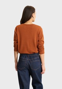 Maglione leggero di colore ruggine, a maglia, con maniche lunghe, scollatura rotonda e vestibilità casual, abbinato a jeans a vita alta blu scuro.