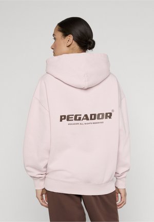 Žena s tmavými vlasy svázanými do drdolu má na sobě světle růžovou mikinu s nápisem "PEGADOR" na zádech, v kombinaci s hnědými kalhotami, otočená zády k fotoaparátu.