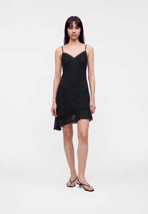 Femme portant une robe noire en dentelle sans manches avec bretelles spaghetti et ourlet asymétrique, associée à des sandales noires à talons, debout sur fond blanc.