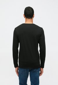 Άνδρας που γυρίζει την πλάτη, φορώντας ένα απλό μαύρο μπλουζάκι με μακριά μανίκια και μπλε τζιν, σε λευκό φόντο.