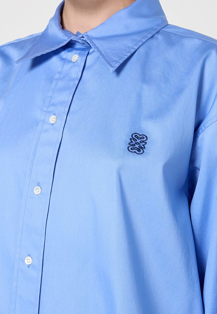 Hellblaues Button-Up-Hemd aus glattem Stoff, mit einem klassischen Kragen und einem kleinen, dunkelblau gestickten Logo auf der linken Brust.