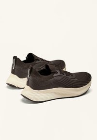Een paar donkerbruine sport Sneakers met textuurgebreide bovenkant, witte gevoerde zolen en trekbandjes aan de hiel op een lichte achtergrond.