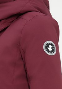 Burgundijas jaka ar gludu tekstūru, kurai ir apaļš pelēks uzlīmju laukums ar pīles logotipu. Audumam ir smalkas šūšanas detaļas.