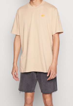 Mężczyzna ubrany w luźny beżowy T-shirt z małym żółtym logo na piersi oraz podniszczone czarne szorty z denimu.