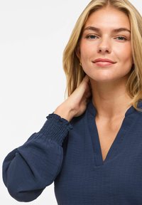 Femme blonde aux yeux bleus portant un chemisier bleu marine aux poignets froncés, touchant son cou et souriant doucement.