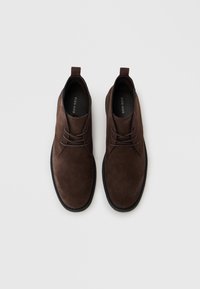 Botas de ante marrón hasta el tobillo con punta redonda y suela de goma negra. Cuentan con cordones y una pestaña en el talón para facilitar el calzado.