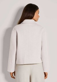 Vrouw met lang donker haar, gekleed in een lichtbeige cropped jack en bijpassende wijde broek, staand met haar gezicht naar weg tegen een effen achtergrond.