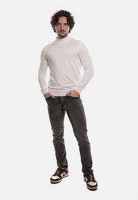 Maglione girocollo bianco in cotone abbinato a jeans grigio scuro e sneakers nere con dettagli bianchi, in piedi davanti a uno sfondo neutro.