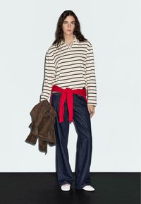 Gestreepte langwerpige shirt in roomkleur en bruin, rode tailleband, donkerblauwe rechte jeans, bruine jas over de arm, witte schoenen.