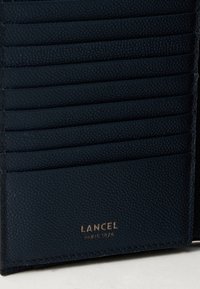 Tumši zilas teksturētas ādas maciņa daļa ar vairākiem karšu slotiem un zelta izspiestu "LANCEL Paris 1876" logotipu.