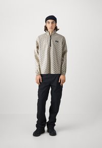Beige patterned pullover with a chevron design, featuring a quarter zip and a small logo patch. Worn with dark cargo pants and black shoes.