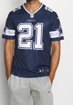Mand iført en navyblå Dallas Cowboys-trøje med nummer 21 og grå Nike-sweatpants, stående med hænderne i lommerne foran en ensfarvet baggrund.