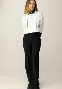 Camicia bianca corta con colletto nero e bottoni dorati, abbinata a pantaloni neri a vita alta con tasche laterali e vestibilità sartoriale.