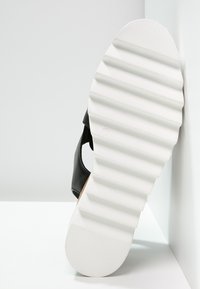 Sandale noire avec une tige en cuir lisse. La semelle en caoutchouc blanche présente un design texturé et strié pour une meilleure adhérence et stabilité.