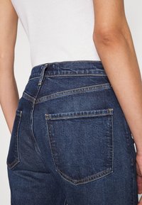 Mörkblå denimjeans med hög midja, som har två bakfickor och synliga söm detaljer. Slät textur, klassisk femficksdesign.