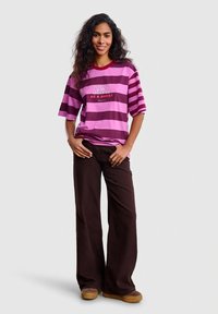 Kobieta z długimi kręconymi włosami stoi uśmiechnięta, ubrana w różowo-fioletowy pasiasty T-shirt oraz ciemne, szerokie spodnie i brązowe buty.