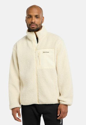 Mann trägt eine cremefarbene Fleecejacke mit Reißverschluss vorne und einer Brusttasche mit der Aufschrift "bruno banani", kombiniert mit schwarzen Hosen.