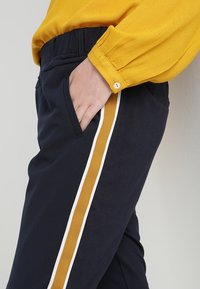 Pantalons navy avec poches latérales présentant des rayures verticales dorées et blanches. Le tissu est lisse avec une taille élastique et un chemisier texturé couleur moutarde.