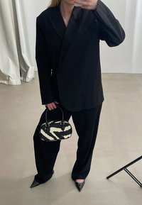 Musta ylipitkä bleiseri, jossa on rakenteelliset hartiat, yhdistettynä leveälahkeisiin mustiin housuihin. Pitää kiinni seepraharmain laukusta ja käyttää kärkipäisiä mustia kenkiä.