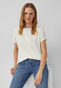 Witte, gestructureerde, kortempers top met een ronde halslijn, gecombineerd met blauwe jeans. Model poseert casual tegen een neutrale achtergrond.