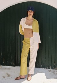 Lichte, twee-kleurige outfit bestaande uit een gele en crème blouse, bijpassende gele broek en een donkerblauwe pet. Eenvoudige sandalen maken de look compleet.