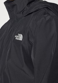 Černá voděodolná bunda s vysokým límcem, předním zipem a bílým logem "The North Face" na levé části hrudi. Hladká textura.