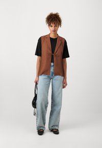 Een bruin linnen vest met een voorknoop, gecombineerd met een zwarte t-shirt, lichtblauwe wijde jeans en een kleine zwarte handtas.